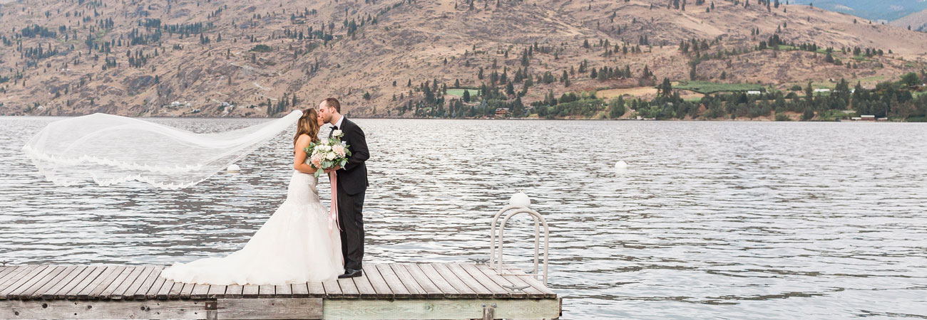 Newleds in wedding attire share a kiss standing upon a dock on the shore of Kelly's Resort